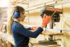 Profile view of a young female woodworker using a drill press on a wood board in a woodshop