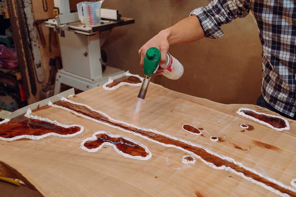 Carpenter using a blow torch to remove air bubbles from epoxy resin on a wooden river table using a blow torch